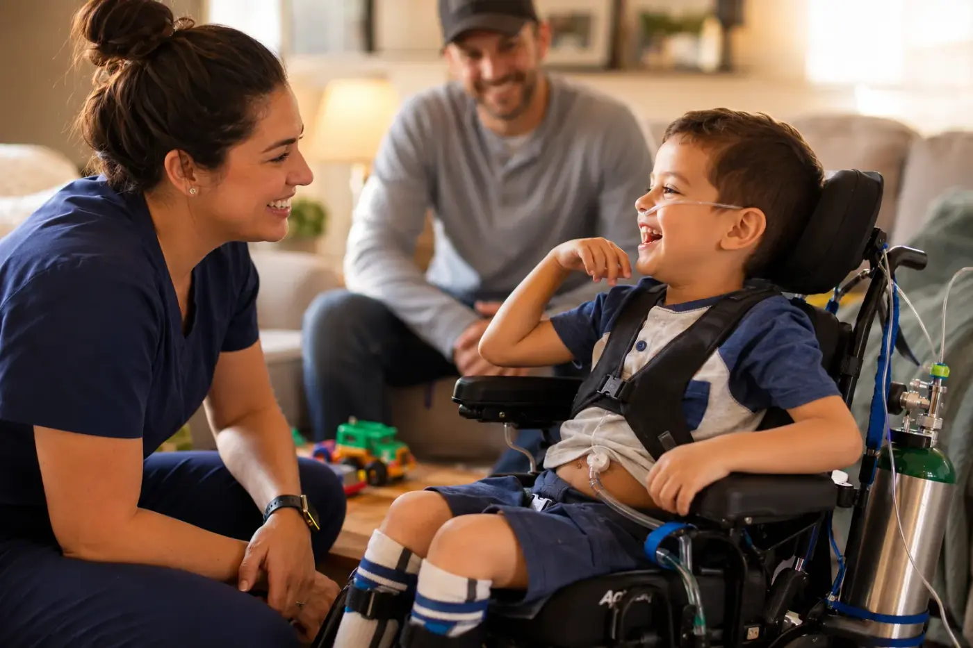 A nurse in navy scrubs kneels beside a smiling boy in a power wheelchair with a nasal cannula, his father smiling behind them.