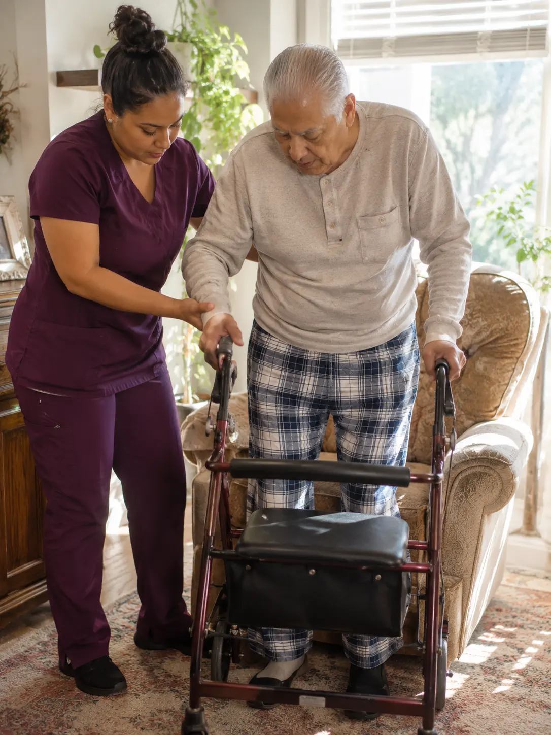 A caregiver in burgundy scrubs steadies an older man as he stands at his walker in his living room.